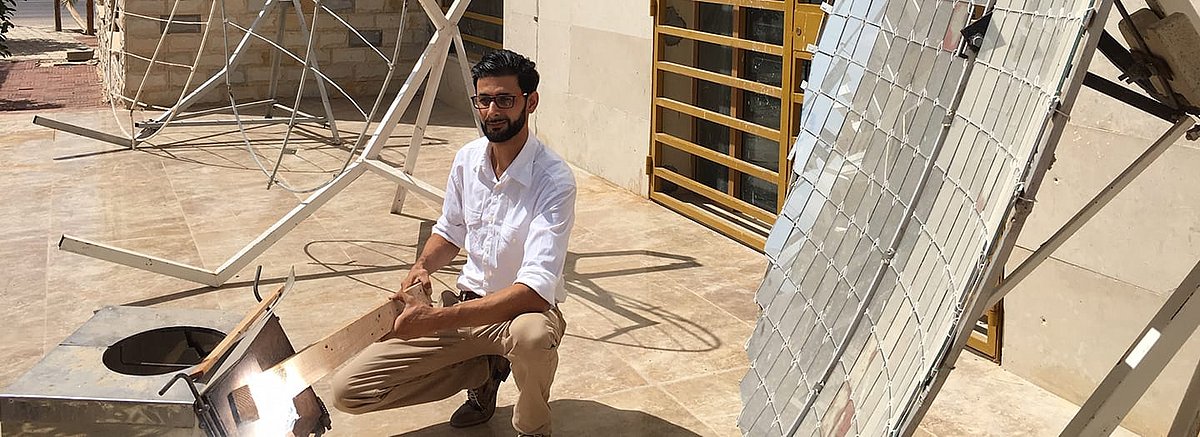 Abdallah Khenissi kniet auf dem Boden und schaut in die Kamera. In der Hand hält er einen Balken aus Holz. Um ihn herum stehen Konstruktionen aus Metall sowie sein Solarkonzentrator.