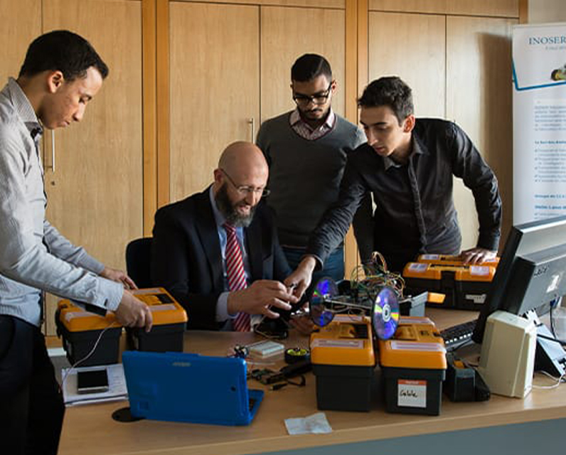 Farid El-Houzia sitzt an einem Schreibtisch. Auf dem Tisch stehen mehrere Boxen und ein Roboter. Um den Tisch herum stehen weitere Mitarbeiter. 