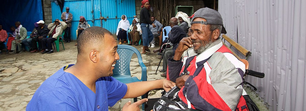 Ein älterer Mann sitzt auf einem Stuhl. Surafel Mengistu kniet ihm gegenüber. Beide lachen freundlich. Im Hintergrund sitzen weitere Menschen auf Stühlen.