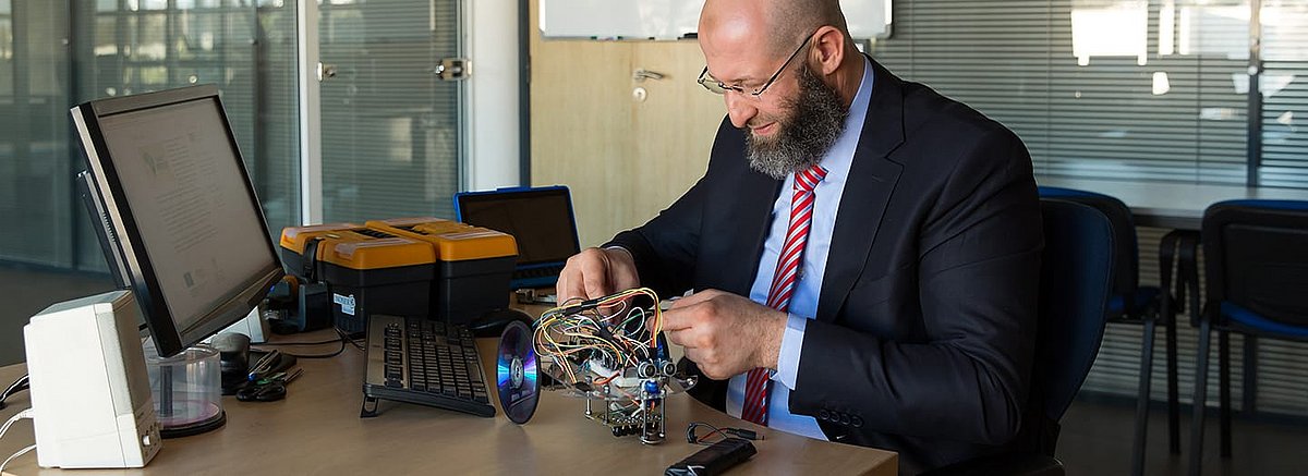 Farid El-Houzia sitzt an einem Schreibtisch und bastelt an einem Roboter, aus dem viele Kabel hervorragen. Er lächelt und schaut gebannt auf die Technik.