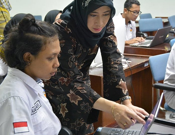 Eine Frau sitzt vor einem Laptop. Neben ihr steht eine Frau und zeigt auf den Bildschirm des Laptops.