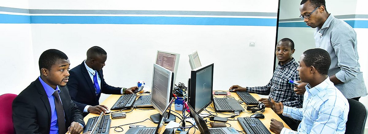 Romeo Nwansi und vier Mitarbeiter befinden sich in einem Büro. Die Mitarbeiter sitzen an Schreibtischen vor ihren Computern. Sie wirken gestresst.