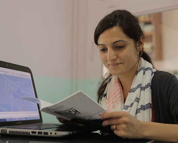 Apekshya Dhungel sitzt an einem Tisch. In der Hand hält sie ein Heft, in dem sie liest. Neben ihr steht ein Laptop auf dem Tisch.