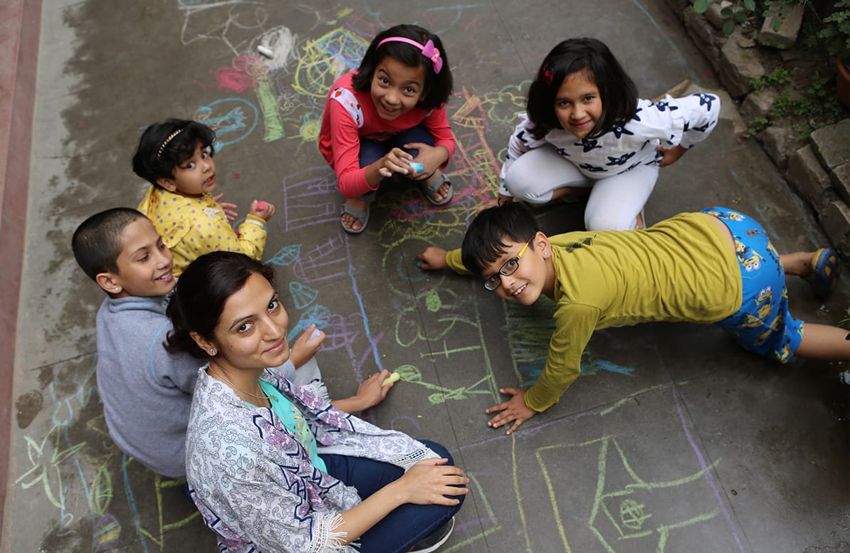 Apekshya Dhungel sitzt auf dem Boden und schaut nach oben in die Kamera. Um sie herum sitzen mehrere Kinder und malen mit Kreide auf den Boden.