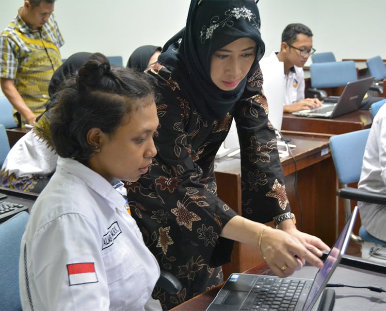 Eine Frau sitzt vor einem Laptop. Neben ihr steht eine Frau und zeigt auf den Bildschirm des Laptops.