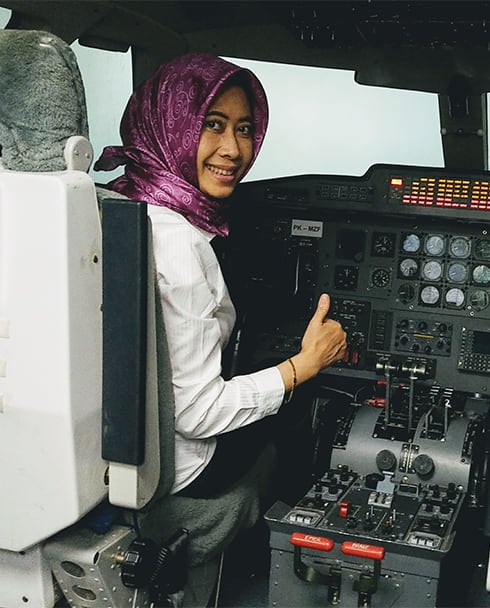 Eine Kopftuch tragende Frau sitzt im Cockpit eines Flugzeuges. Sie zeigt Daumen hoch in die Kamera und lächelt.