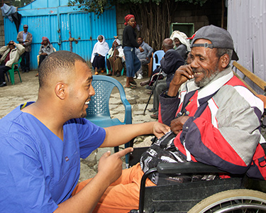Ein älterer Mann sitzt auf einem Stuhl. Surafel Mengistu kniet ihm gegenüber. Beide lachen freundlich. Im Hintergrund sitzen weitere Menschen auf Stühlen.