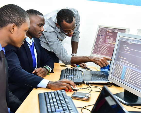 Romeo Nwansi und zwei Mitarbeiter sitzen an einem Schreibtisch vor zwei Computer. Nwansi zeigt etwas auf einem der Bildschirme.