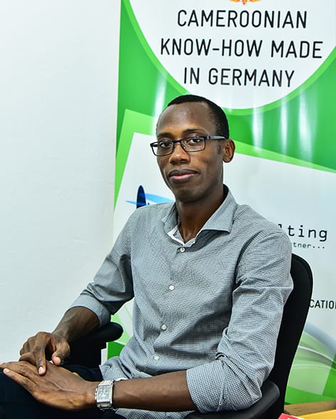 Romeo Nwansi sitzt auf einem Bürostuhl vor einem grün-weißen Banner und lächelt in die Kamera.