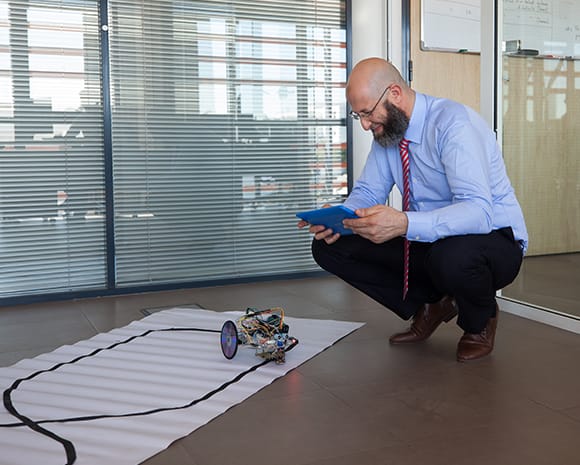 Farid El-Houzia hält ein Tablet in beiden Händen. Mit dem Tablet steuert er einen Roboter, der auf dem Boden steht.