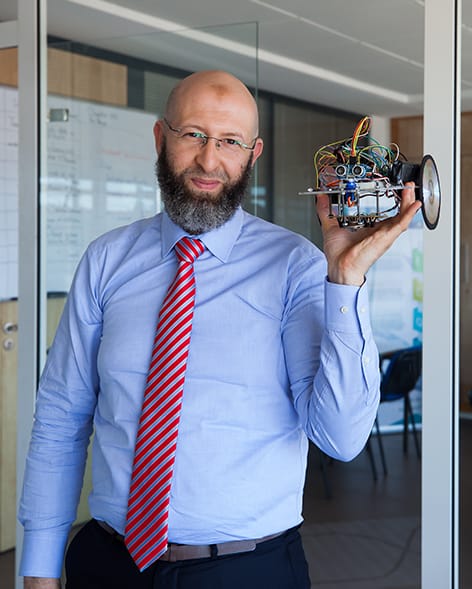 Farid El-Houzia hält in einer Hand einen zusammengebastelten Roboter, aus dem viele Kabel hervorragen, stolz in die Kamera.