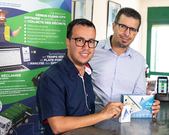 Salim Benaissa (rechts) und sein Geschäftspartner Aziz Ahnyne stehen vor einem Banner ihrer Firma. Vor ihnen liegt ihr GPS-Trackingsystem auf einem Tisch. 