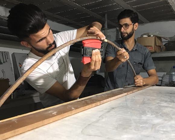 Abdallah Khenissi und ein Mitarbeiter befinden sich in einer Werkstatt. Zusammen arbeiten sie an einem runden Holzrahmen. 