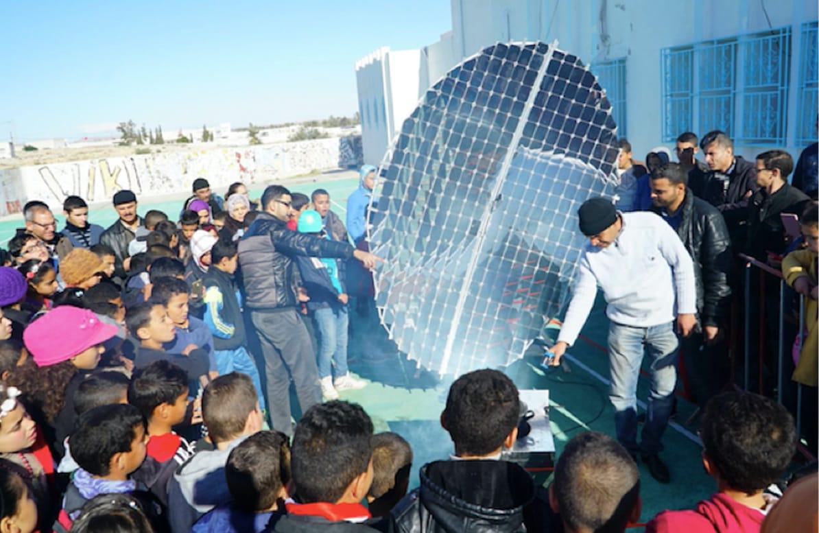 Abdallah Khenissi zeigt einer Gruppe von Menschen seinen Solarkonzentrator. Die Gruppe steht in einem großen Kreis um das Gerät herum.